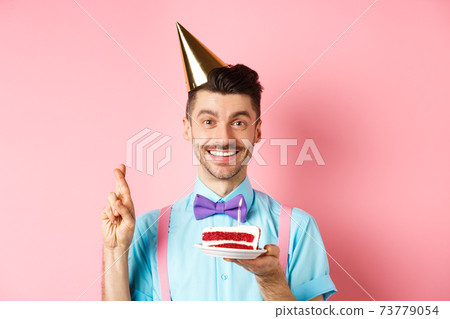 Holidays and celebration concept. Happy young man enjoying birthday party, wearing cone hat and cross fingers, making wish on bday cake with candle, pink background 73779054