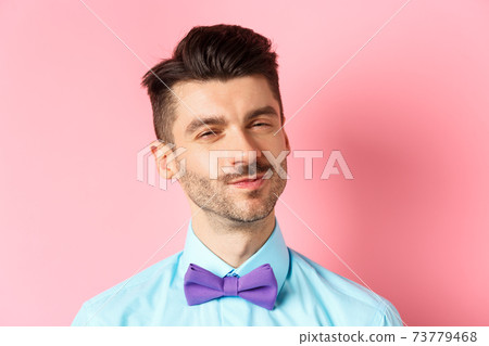 Close-up of smiling young guy with moustache, squinting intrigued, listening to something interesting, standing in bow-tie on pink background 73779468