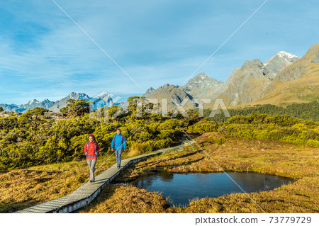 New Zealand Hiking. Young hiking couple walking on trail at Routeburn Track during sunny day. Hikers are carrying backpacks while tramping Key Summit Track in Fiordland National Park in New Zealand 73779729