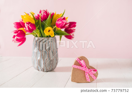 Craft heart shape gift box with satin ribbon bow and spring bouquet of pink and yellow tulip flowers in a vase on the white wooden table and pink background. Greeting card mockup. Copy space. Craft heart shape gift box with satin ribbon bow and spring bouquet of pink and yellow tulip flowers in a vase on the white wooden table and pink background. Greeting card mockup. Copy space. 73779752