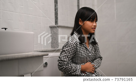 Asian girl holding her belly and.suffering from stomachache while sitting in toilet at home. Asian girl holding her belly and.suffering from stomachache while sitting in toilet at home. 73791165