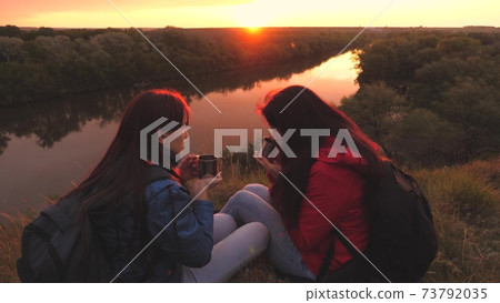 Female travelers sit high on a mountain by the river at sunset and drink tea from a mug. Go camping with a friend on the weekend. Tourist exploration of the area of nature. Free yourself from everyday 73792035