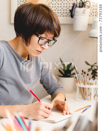 Woman with short hair cut is drawing in notebook. Calming hobby, anti stress leisure. Artist at work. Cozy workplace. Woman with short hair cut is drawing in notebook. Calming hobby, anti stress leisure. Artist at work. Cozy workplace. 73793388