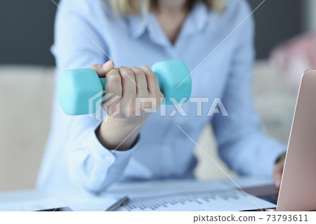 Woman sits on laptop and holds blue dumbbell in her hand Woman sits on laptop and holds blue dumbbell in her hand 73793611