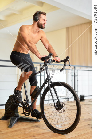 Cheerful athletic man riding stationery bicycle and listening to music Cheerful athletic man riding stationery bicycle and listening to music 73801094
