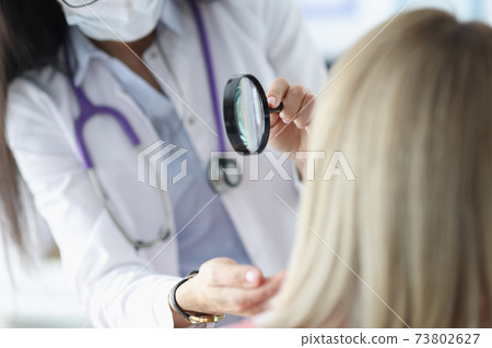 Dermatologist examines patient's face through magnifying glass Dermatologist examines patient's face through magnifying glass 73802627
