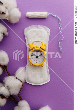 Menstrual pad, cotton tampon and yellow alarm clock on lilac background. Female's menstrual cycle concept. Vertical image 73803610
