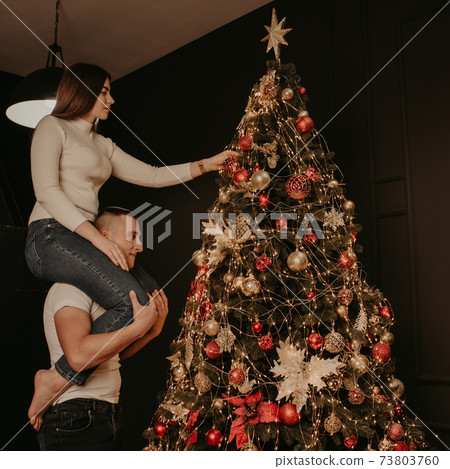 young woman sits on back shoulders of man and decorates a Christmas tree young woman sits on back shoulders of man and decorates a Christmas tree 73803760