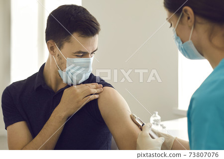 Man in face mask looking at syringe while getting Covid-19 vaccine at the clinic 73807076