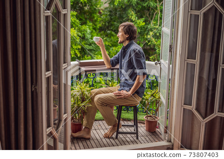man stands on a heritage-style balcony enjoying his morning coffee. A man in a hotel in Europe or Asia as tourism recovers from a pandemic. Tourism has recovered thanks to vaccinations. Woman man stands on a heritage-style balcony enjoying his morning coffee. A man in a hotel in Europe or Asia as tourism recovers from a pandemic. Tourism has recovered thanks to vaccinations. Woman 73807403
