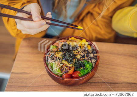 Woman eating Raw Organic Poke Bowl with Rice and Veggies close-up on the table. Top view from above horizontal 73807867