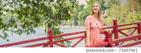 BANNER, LONG FORMAT Caucasian woman traveler on background of Red Bridge in public park garden with trees and reflection in the middle of Hoan Kiem Lake in Downtown Hanoi. Vietnam reopens after 73807961
