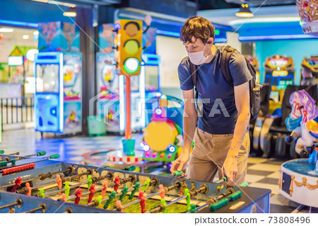 A young man wearing a medical mask during COVID-19 coronavirus playing table football indoors against a friend A young man wearing a medical mask during COVID-19 coronavirus playing table football indoors against a friend 73808496