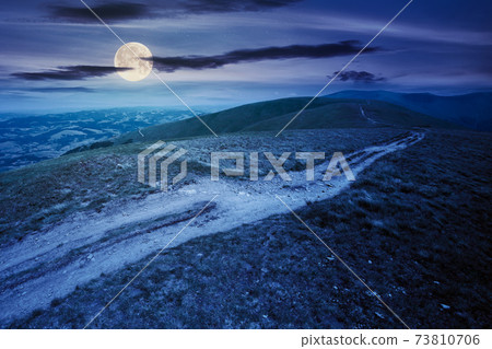 mountain road through grassy meadow at night. wonderful summer adventure in full moon light. clouds on the blue sky 73810706