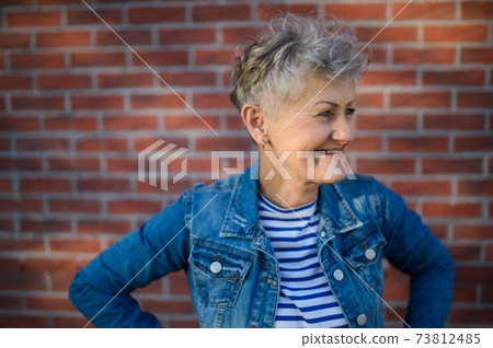 Senior woman standing outdoors against brick wall background. Copy space. Senior woman standing outdoors against brick wall background. Copy space. 73812485