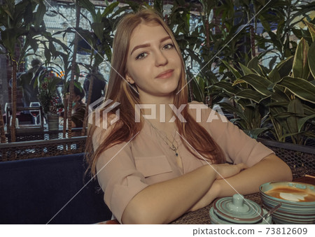 Blond girl in cafeteria near the potted plants with coffee in front of her looking at camera. Cafe city lifestyle woman drinking coffee. 73812609