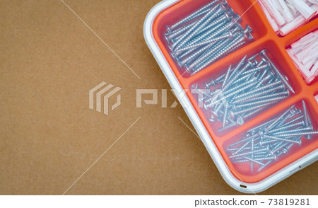 Steel silver screws and wall plug in orange plastic box. Top view tool box on brown paper box background with copy space. Set of screws and plastic plug for drilling wall and installation things. Steel silver screws and wall plug in orange plastic box. Top view tool box on brown paper box background with copy space. Set of screws and plastic plug for drilling wall and installation things. 73819281