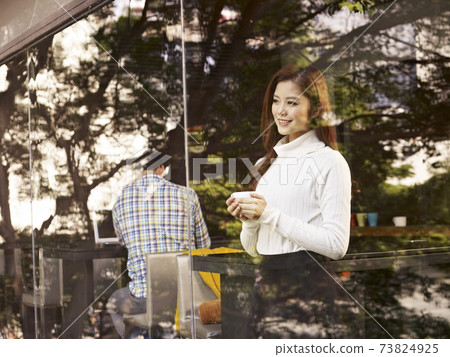 young asian woman standing in front of windows with coffee cup in hand in cafe. 73824925
