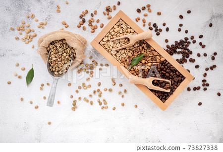 Green and brown decaf unroasted and dark roasted coffee beans in wooden box with scoops setup on white concrete background. Green and brown decaf unroasted and dark roasted coffee beans in wooden box with scoops setup on white concrete background. 73827338