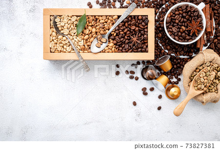 Green and brown decaf unroasted and dark roasted coffee beans in wooden box with scoops setup on white concrete background. 73827381
