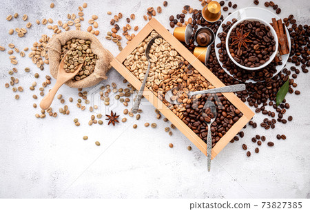 Green and brown decaf unroasted and dark roasted coffee beans in wooden box with scoops setup on white concrete background. 73827385