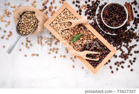 Green and brown decaf unroasted and dark roasted coffee beans in wooden box with scoops setup on white concrete background. Green and brown decaf unroasted and dark roasted coffee beans in wooden box with scoops setup on white concrete background. 73827387