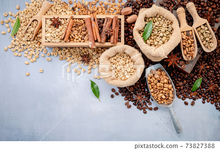 Green and brown decaf unroasted and dark roasted coffee beans in hemp sack bags with scoops setup on white concrete background. Green and brown decaf unroasted and dark roasted coffee beans in hemp sack bags with scoops setup on white concrete background. 73827388