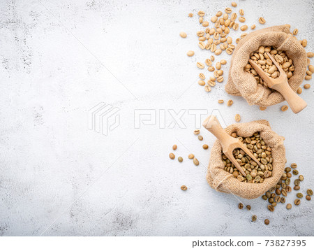 Green and brown decaf unroasted coffee beans in hemp sack bags with scoops setup on white concrete background. Green and brown decaf unroasted coffee beans in hemp sack bags with scoops setup on white concrete background. 73827395