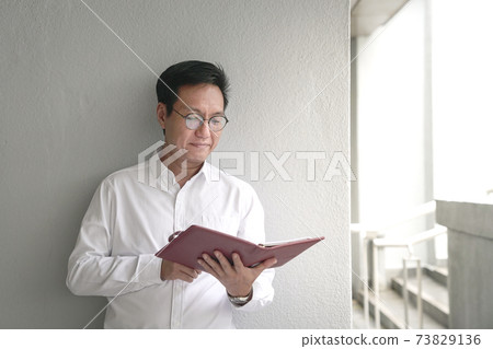 Businessman leaning against white wall, reading at his appointment book. 73829136