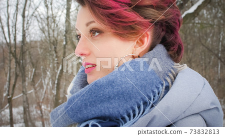 Attractive young woman with perfect skin and makeup with dark pink hair is possing on winter park background wearing blue scarf and silver earrings 73832813