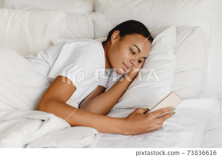 African american female laying relaxing on bed holding up smart phone. African american female laying relaxing on bed holding up smart phone. 73836616