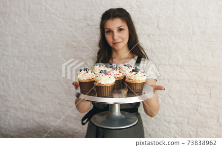 The cook girl holds a tray with beautiful cupcakes decorated with cream and berries. 73838962