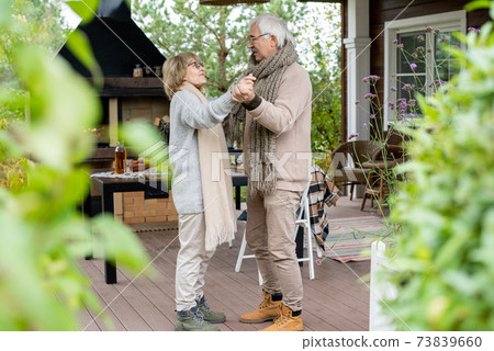Amorous senior couple in warm casualwear dancing on wooden floor of patio 73839660