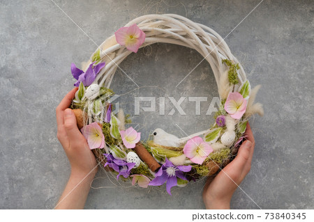 Female hands hold an Easter wreath with flowers and a bird on a gray Female hands hold an Easter wreath with flowers and a bird on a gray 73840345