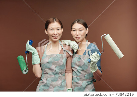 Happy young Asian female twins in aprons and gloves holding paint rollers Happy young Asian female twins in aprons and gloves holding paint rollers 73842219