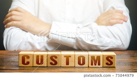 Man with folded hands on his torso and blocks with word Customs. Customs border control. Tariffs and taxes on the import of goods and products. Imports monitoring. Duties. Transportation and transit. Man with folded hands on his torso and blocks with word Customs. Customs border control. Tariffs and taxes on the import of goods and products. Imports monitoring. Duties. Transportation and transit. 73844979