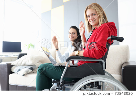 Woman in wheelchair with laptop on her lap waving in greeting with her girlfriend 73848020