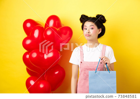 Happy Valentines day. Stylish asian single girl buying herself gift, holding shopping bag and looking unbothered at camera, standing over yellow background 73848581