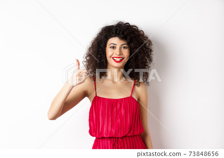 Cheerful woman with red lips and elegant dress, showing thumb up and smiling, recommending product, standing over white background 73848656