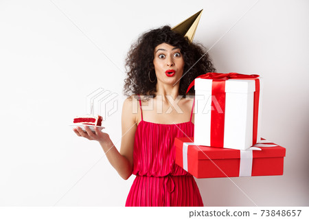 Funny lady in red dress and party hat, celebrating birthday, holding b-day presents and cake with candle, looking amused at camera, standing over white background 73848657