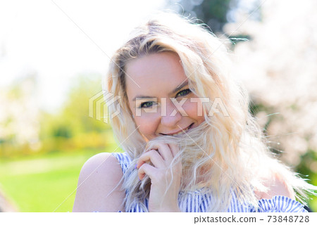 Waist up portrait of young smiling woman with blond hair near blossoming plum tree 73848728
