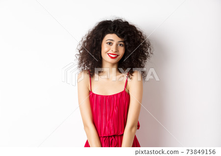 Cute modest lady in red dress and makeup, smiling and blushing, looking at camera, standing over white background Cute modest lady in red dress and makeup, smiling and blushing, looking at camera, standing over white background 73849156