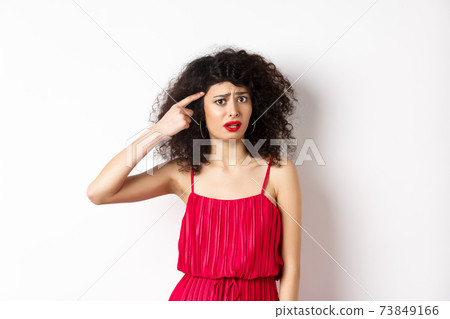 Shocked and confused caucasian woman in red dress scolding someone crazy, pointing finger at head and frowning, standing over white background 73849166