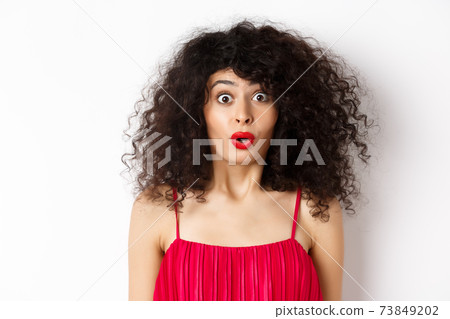 Close-up of surprised caucasian woman with red lips and curly hairstyle, saying wow and looking amazed, standing in red dress over white background 73849202