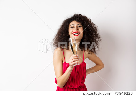 Cheerful elegant woman with dark curly hair, wearing party dress, celebrating anniversary on valentines day, drinking champagne from glass and smiling at camera, white background Cheerful elegant woman with dark curly hair, wearing party dress, celebrating anniversary on valentines day, drinking champagne from glass and smiling at camera, white background 73849226