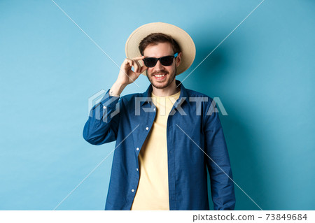 Happy smiling guy going on summer vacation, wearing straw hat and black sunglasses, standing on blue background 73849684