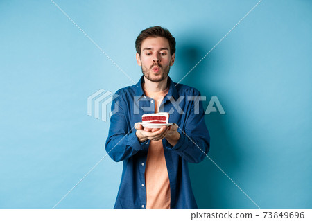 Celebration and holiday concept. Handsome young man blowing candle on birthday cake and making wish, standing on blue background 73849696