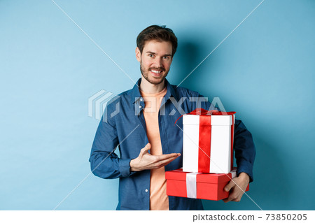 Young man in casual clothes buying romantic gifts on Valentines day, pointing at presents boxes and smiling, standing over blue background 73850205