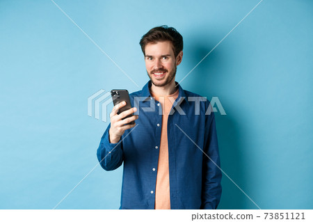 Attractive caucasian man using mobile phone, looking happy and smiling at camera, standing on blue background 73851121
