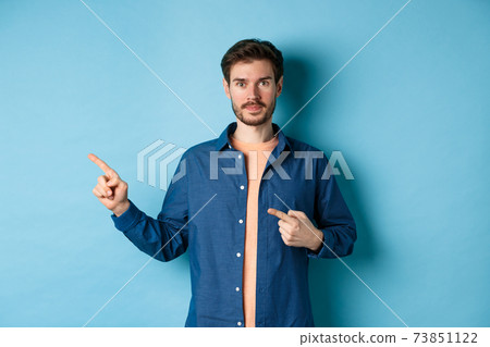 Handsome young man in shirt, pointing fingers left at promo, showing logo, standing on blue background Handsome young man in shirt, pointing fingers left at promo, showing logo, standing on blue background 73851122
